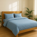 A misty blue linen duvet set with a zipper, displayed on a bed in a room with wooden flooring and a plant in the background.